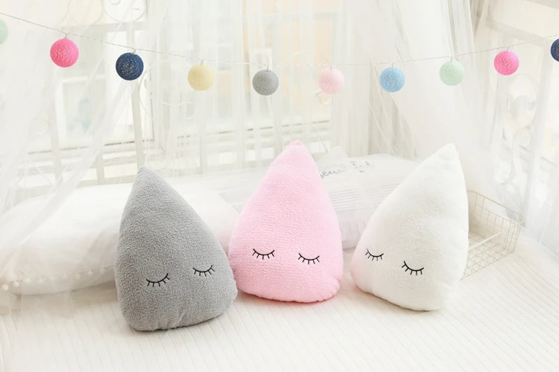 Three cloud-shaped pillows in gray, pink, and white with closed eyes on a light background.
