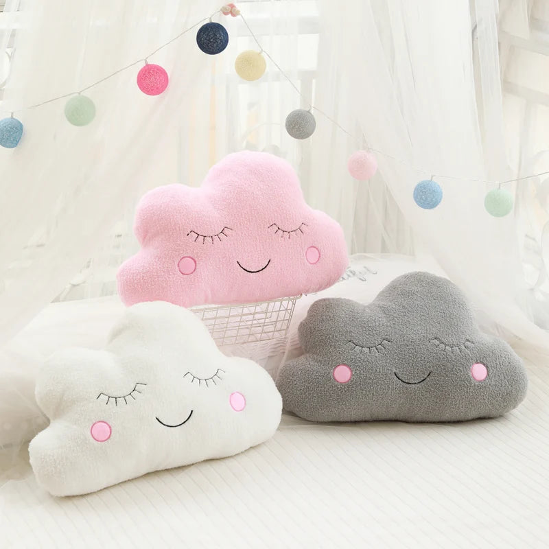 Three cloud-shaped pillows in pink, gray, and white with smiling faces on a light background.