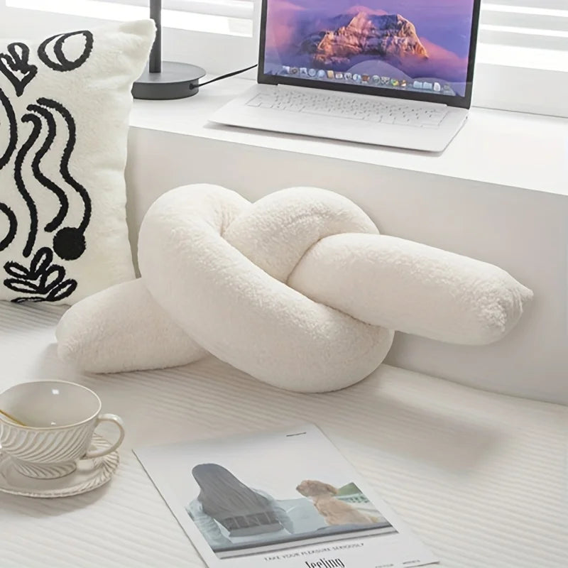 White knot-shaped pillow on a desk with a laptop, cup, and magazine in the background.