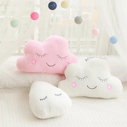 Set of cloud-shaped pillows with smiling faces on a soft surface with colorful polka dot string lights in the background.